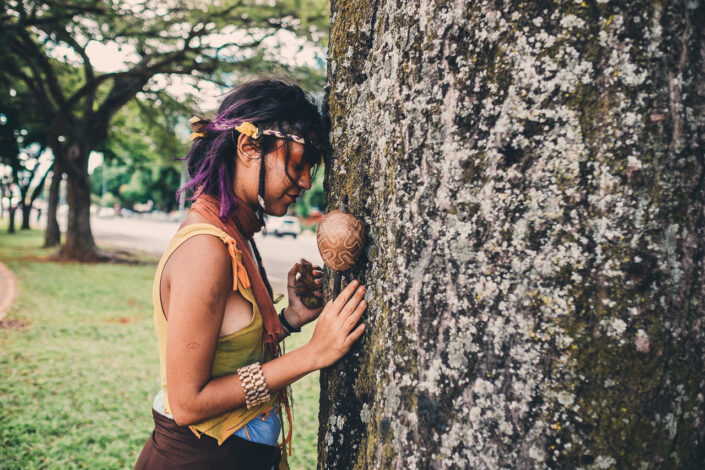 Jeune femme indigène qui embrace un arbre lors de la manifestation sur l'esplanade des ministères avec dans sa main droite une Maracá, l'instrument symbolique de la lutte indigène et dans sa main gauche des fruits de l'urucum, qui fait leur peinture rouge.